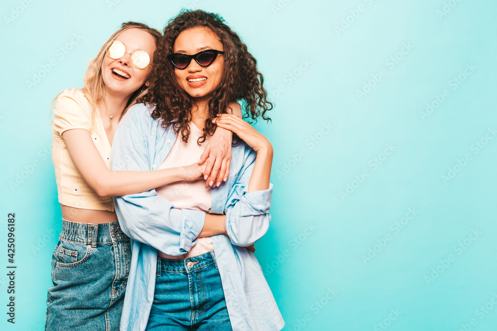 Two young beautiful smiling international hipster female in trendy summer clothes. Sexy carefree women posing near blue wall in studio. Positive models having fun. Concept of friendship
