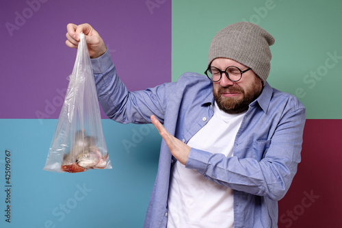 Man holds a smelly, spoiled fish in a transparent bag, closes eyes because of the unpleasant smell and makes a stop gesture. 