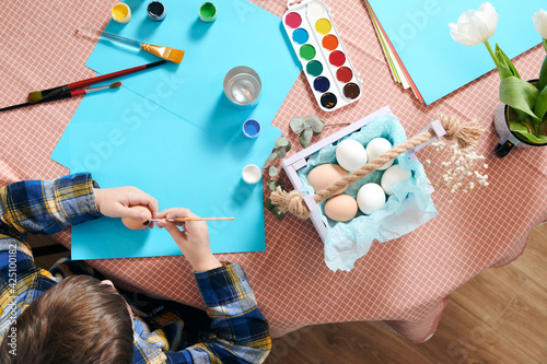 Little child paints easter eggs on a table with white flower
and paints for painting. Easter concept. 