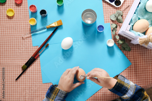 Child hand painting easter egg with paintbrush. Easter eggs on a table with white flower
and paints for painting. Easter concept. 