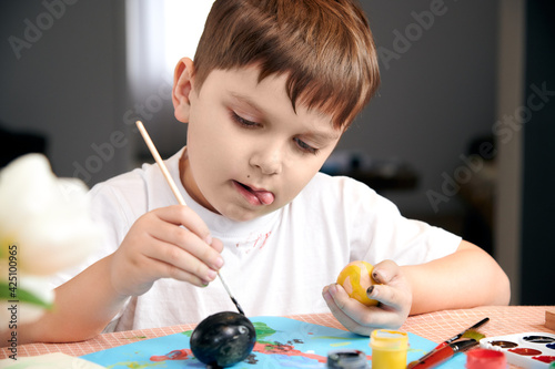 child paints Easter eggs on a colored table. the child draws with his tongue out.