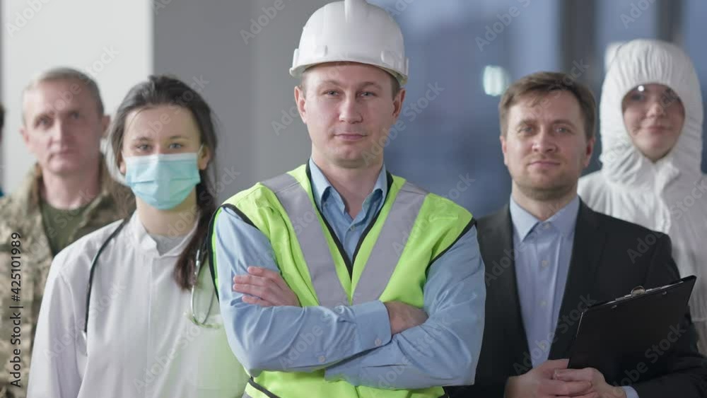 Confident smiling male builder looking at camera crossing hands standing indoors with group of people of different professions. Portrait of Caucasian man in hard hat posing. Construction industry Confident smiling male builder looking at camera crossing hands standing indoors with group of people of different professions. Portrait of Caucasian man in hard hat posing. Construction industry