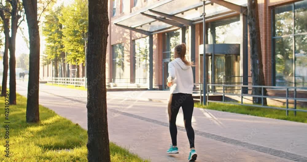 Rear view of athletic female sportwoman running along modern office buildings. Adult Caucasian lady doing sports, working out, jogging in urban city street. Healthy and wealthy lifestyle concept.