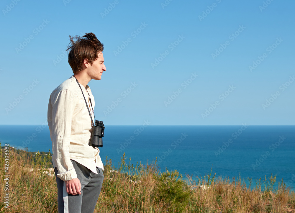 A side view of an attractive young man with binoculars hanging around his neck in front of lovely sea scenery on a windy day