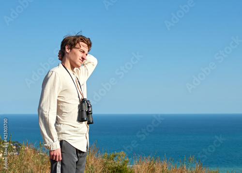 A portrait of a tired young man with binoculars hanging around his neck standing in front of lovely sea scenery looking afar © Érik Glez.