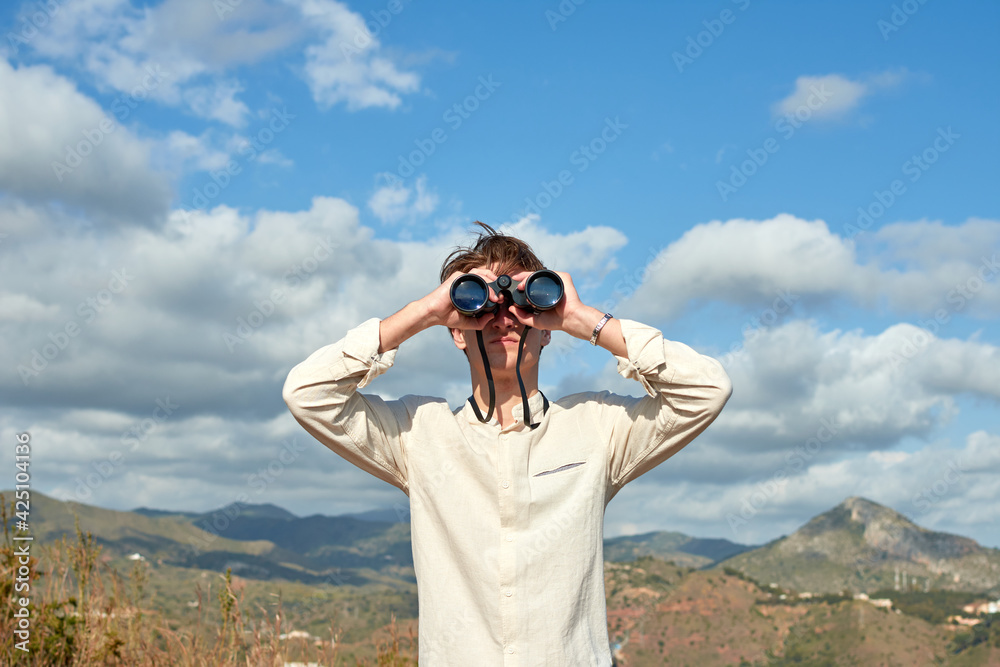 A young caucasian male looking at a view with binoculars under a sky with clouds