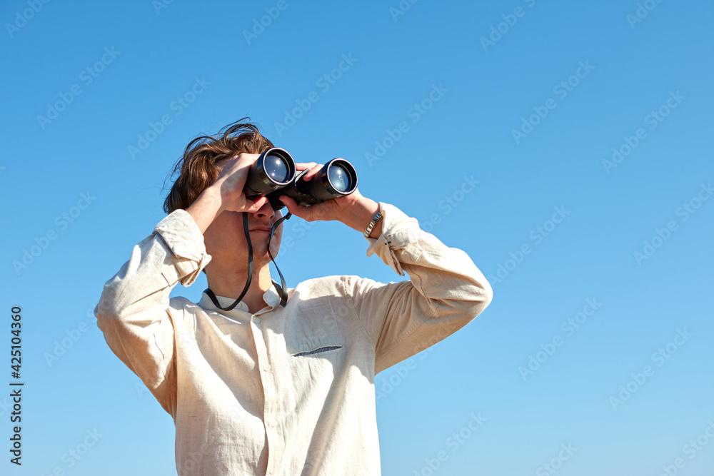 A young caucasian male looking at a view with binoculars under the bright blue sky