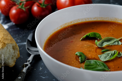 Close-up of a bowl with tomato soup and basil, next to it a baguette.