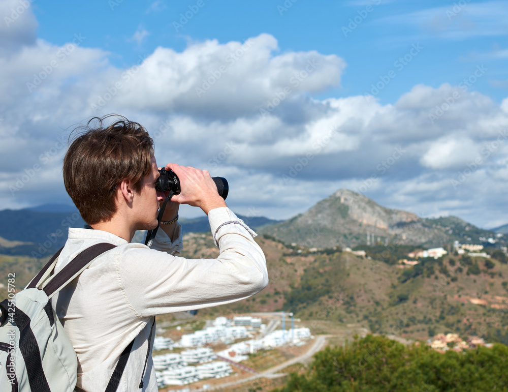 A Shallow focus shot of a young traveler from Spain in a white shirt looking over mountains through binoculars A Shallow focus shot of a young traveler from Spain in a white shirt looking over mountains through binoculars