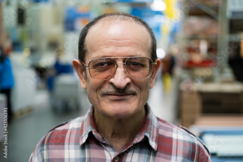 A senior man with a mustache, glasses, and a plaid shirt looks at the camera.