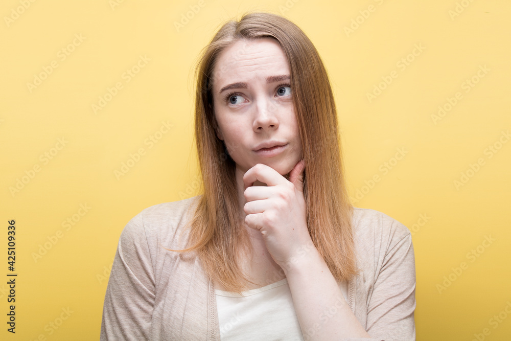 A blonde girl on a light background looks at the camera and holds her hand at her chin with doubt. Concept - the girl doubts. A blonde girl on a light background looks at the camera and holds her hand at her chin with doubt. Concept - the girl doubts.