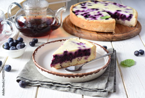 Delicious blueberry cake and a slice of blueberry cake on a white plate. A piece of berry pie on a nice plate and a cup of hot tea on a white table.