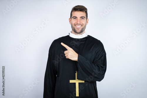 Young hispanic man wearing priest uniform standing over white background smiling and pointing with hand and finger to the side