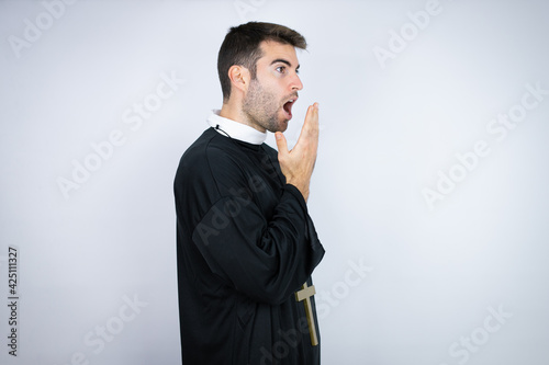 Young hispanic man wearing priest uniform standing over white background covering mouth with hand, shocked and afraid for mistake. surprised expression