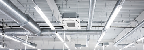 Ceiling mounted cassette type air condition units with other parts of ventilation system (tubes, cables and vents) located inside commercial hall with hanging lights and other construction parts.