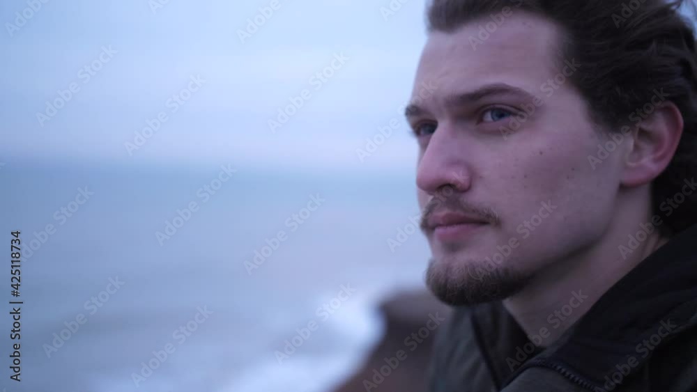 Attractive blue-eyed young man with beard and curly hair lone portrait on a windy cliff above dark sea. Looking worried, He look up on troubled water and cloudy sky with a hope at late autumn Attractive blue-eyed young man with beard and curly hair lone portrait on a windy cliff above dark sea. Looking worried, He look up on troubled water and cloudy sky with a hope at late autumn
