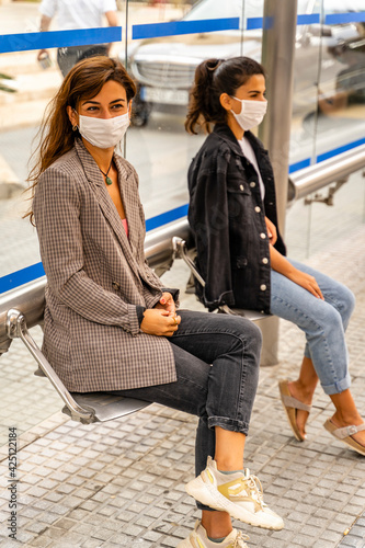 Two Girls Sit in Bus Stop, Wearing Face Mask - Protection Against Covid in Public Place