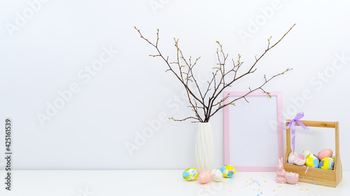 Trendy colorful Easter composition with hand painted eggs in yellow and grey colors in basket, geometric vase, confetti, twigs, pink Easter bunny. Blank photo frame, mockup with copy space for text.