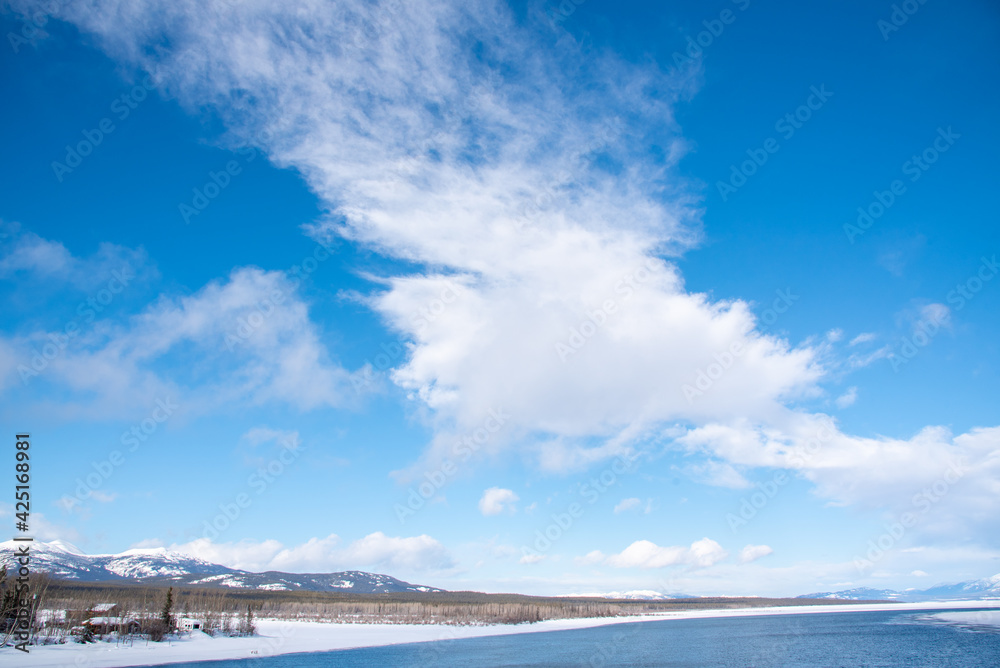 A blue sky bright day with partial clouds and running large river with snow capped mountains and snowy landscape surrounding in Tagish, Yukon Territory, Northern Canada. 