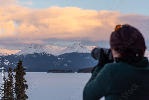 A man with brown hair taking photos of a mountain view scenic spot in northern Canada during winter time March month with snow covered lake, frozen surroundings and mountain peaks.  © Scalia Media