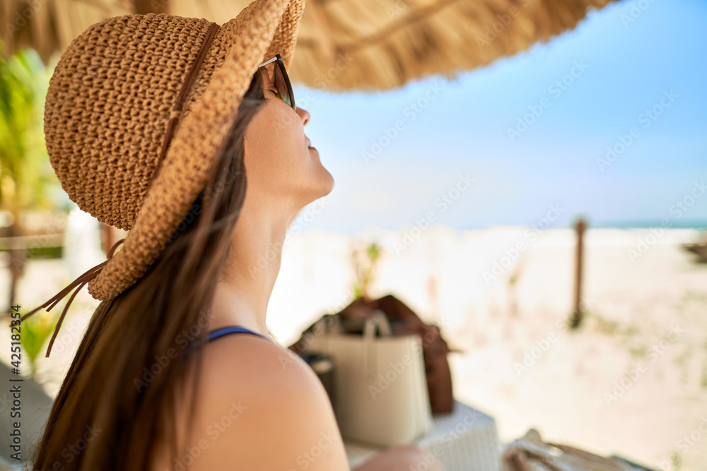 Young woman with a hat in the beach enjoying the ocean breeze under a palm roof