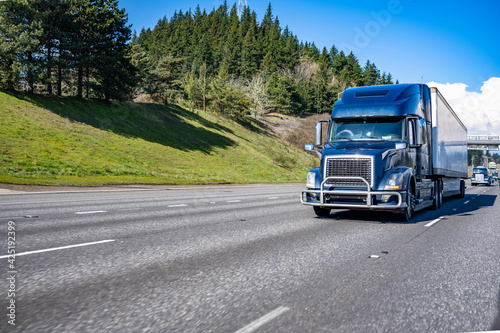 Blue big rig semi truck with pipe grille guard transporting commercial cargo in dry van semi trailer running on the flat straight highway road in front of another track