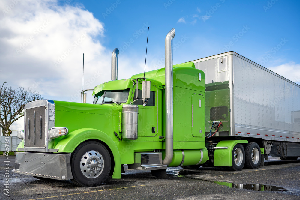 Bright green classic American Idol big rig semi truck tractor with shiny chrome parts and high vertical exhaust pipes and dry van semi trailer standing on the parking lot waiting for the next load Bright green classic American Idol big rig semi truck tractor with shiny chrome parts and high vertical exhaust pipes and dry van semi trailer standing on the parking lot waiting for the next load