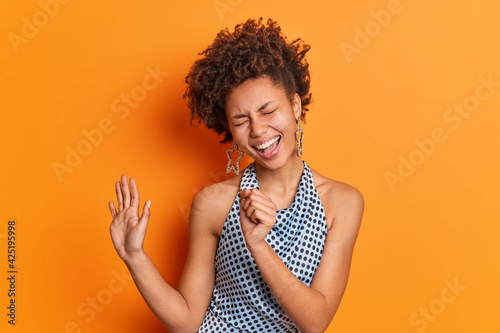 Isolated shot of curly haired woman keeps hand near mouth pretends singing into microphone wears fashionable clothes keeps palm raised isolated over vivid orange background. Stylish lady has fun