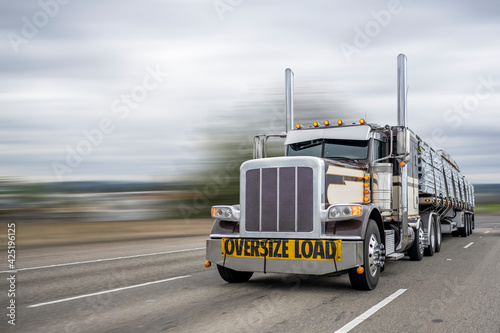 Powerful classic dark big rig semi truck with oversize load sign on the front transporting lumber on the flat bed semi trailer running on the wide highway road
