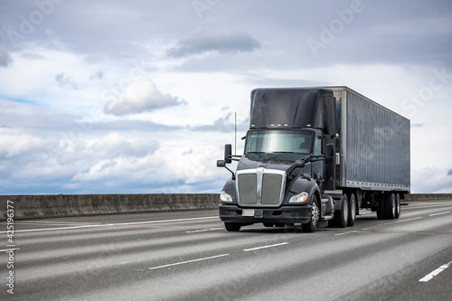 Powerful stylish black big rig day cab semi truck deliver commercial cargo in covered dry van semi trailer driving on the interstate highway road