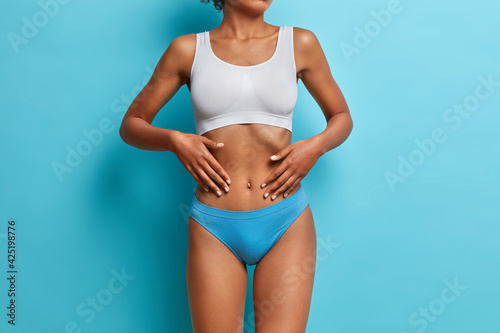 Studio shot of unrecognizable woman has dark skin touches flat stomach poses fit dressed in cropped top and panties isolated over blue background