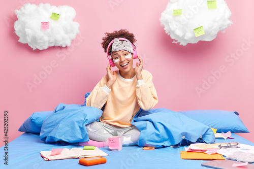 Happy curly teenage girl enjoys lazy day in bed listens music from playlist wears headphones casual pajama sits crossed legs on bed takes break during exam preparation does paperwork at home photo