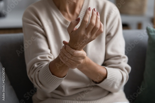 Middle aged elderly woman feeling wrist pain, sitting on couch at home, holding arm. Senior 60s pensioner suffering from rheumatoid arthritis, joints ache, muscle strain, inflammation. Close up photo