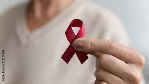 Lady showing hiv or aids awareness symbol, volunteering for charity campaign for prevention immunity disease, cancer, elderly healthcare support. Hand of mature woman holding red ribbon. Close up photo