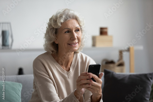 Happy pensive mature lady holding smartphone at home, looking away and smiling. Elderly 60s woman thinking over phone video call for talk to doctor, using online telemedicine app on cell, chatting