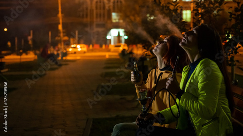 Two caucasian women smoke vape and hookah outdoors at night.
