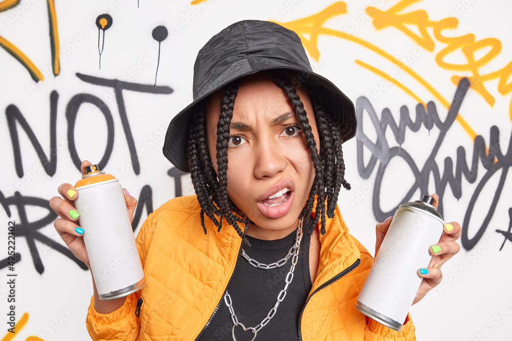 Discontent teenage girl with braids holds aerosol cans for making graffiti dressed in yellow jacket black hat metal chains around neck enjoys urban street liifestyle smirks face. Funky hipster - obrazy, fototapety, plakaty 
