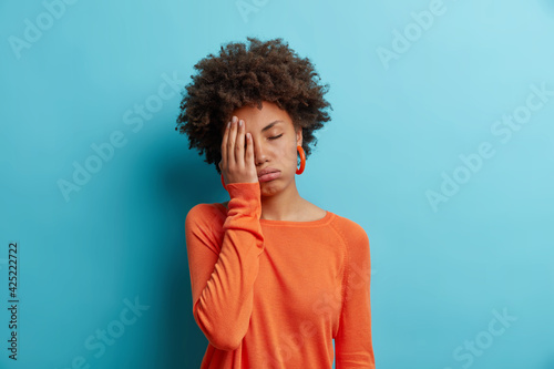 Bored sleepy woman with curly hair makes face palm feels tired or exhausted stands alone closes eyes wears casual orange jumper isolated over blue wall. Attractive female model has gloomy expression