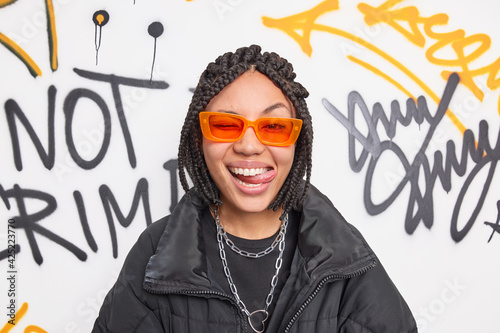 Cheerful fancy teenage girl wears trendy orange suglasses and black jacket sticks out tongue has dreadlocks hairstyle expresses positive emotions poses against graffiti wall. Swag and style concept photo