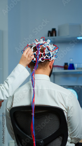 Back view of man patient wearing performant brainwave scanning headset sitting in neurological research laboratory while medical researcher adjusting it, examining nervous system typing on tablet. photo