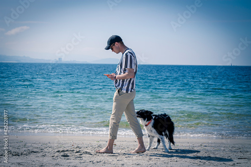 young man walking along the beach shore with his dog using the phone