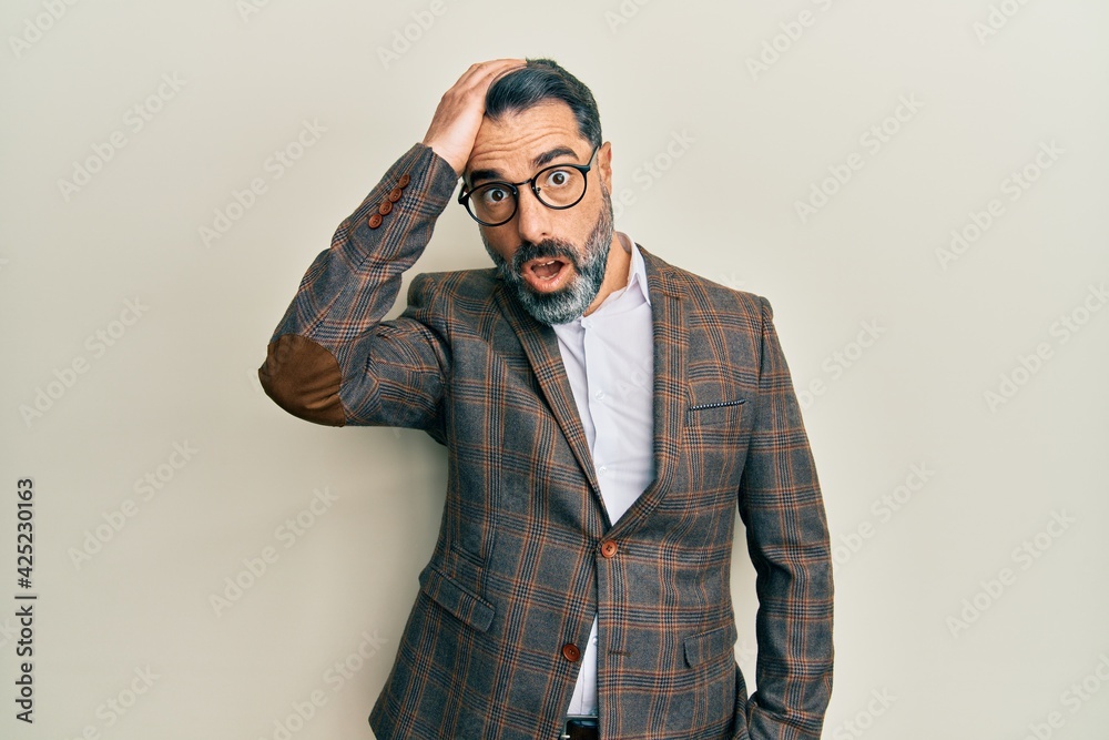 Fototapeta premium Middle age man with beard and grey hair wearing business jacket and glasses surprised with hand on head for mistake, remember error. forgot, bad memory concept.