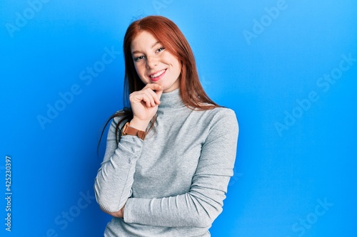 Young red head girl wearing turtleneck sweater smiling looking confident at the camera with crossed arms and hand on chin. thinking positive.