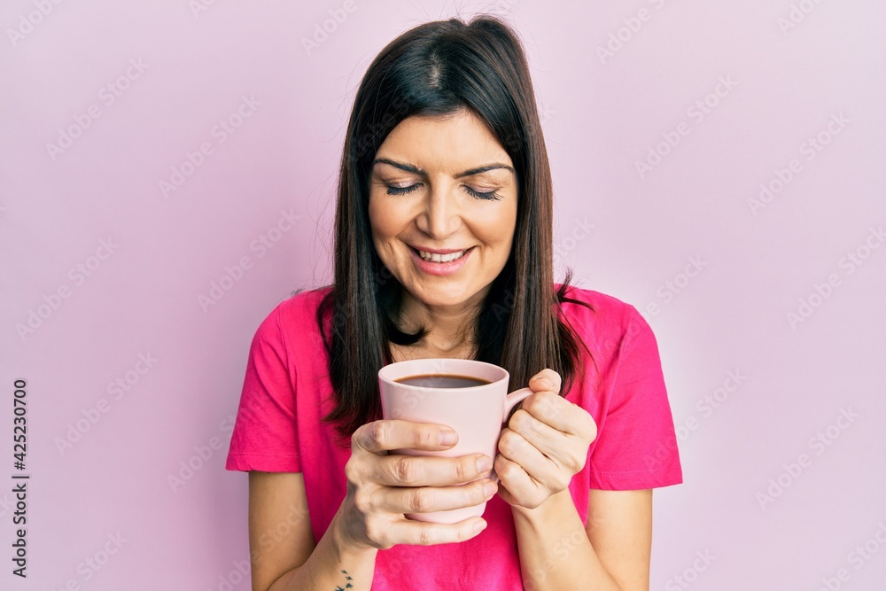Young hispanic woman drinking a cup of coffee smiling happy doing ok sign with hand on eye looking through fingers Young hispanic woman drinking a cup of coffee smiling happy doing ok sign with hand on eye looking through fingers