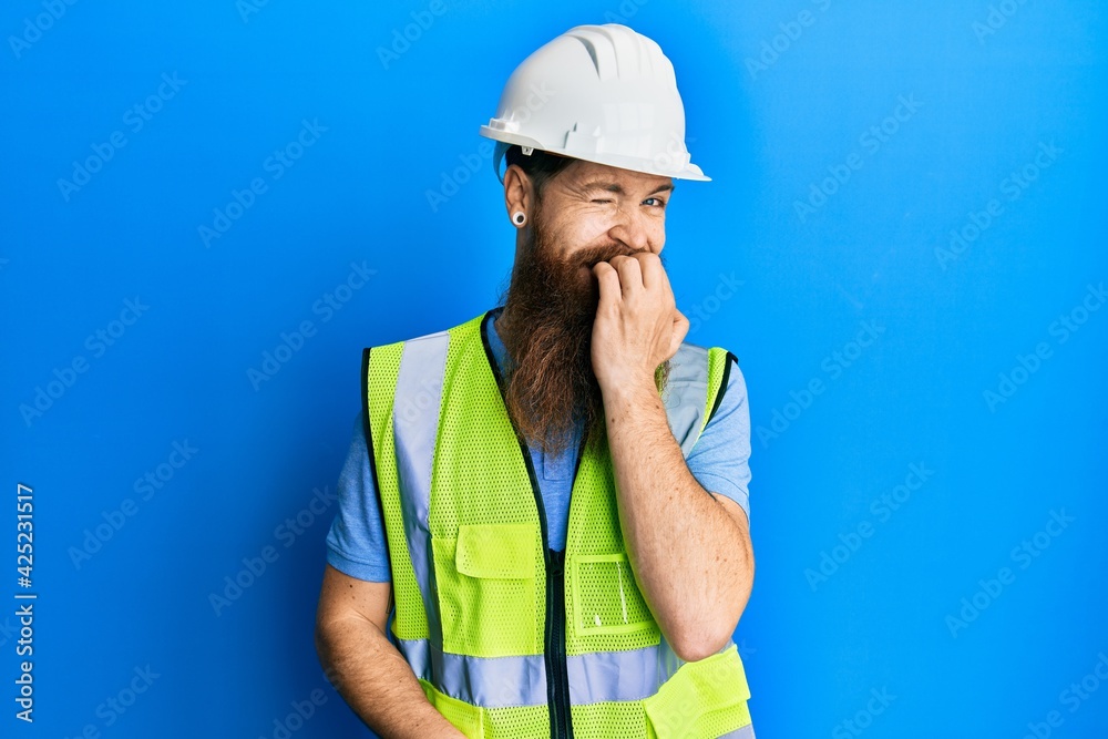 Redhead man with long beard wearing safety helmet and reflective jacket looking stressed and nervous with hands on mouth biting nails. anxiety problem.