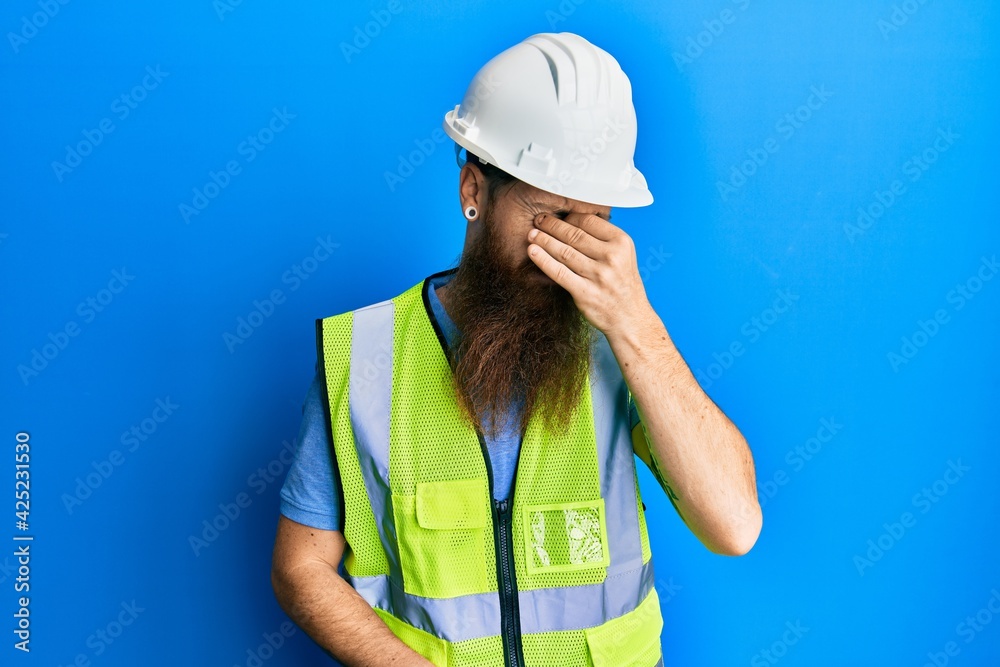 Redhead man with long beard wearing safety helmet and reflective jacket tired rubbing nose and eyes feeling fatigue and headache. stress and frustration concept.