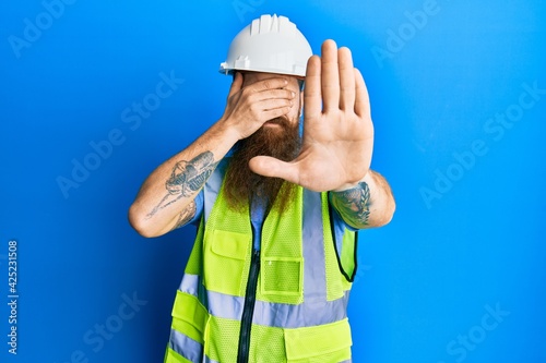 Canvas-taulu Redhead man with long beard wearing safety helmet and reflective jacket covering eyes with hands and doing stop gesture with sad and fear expression