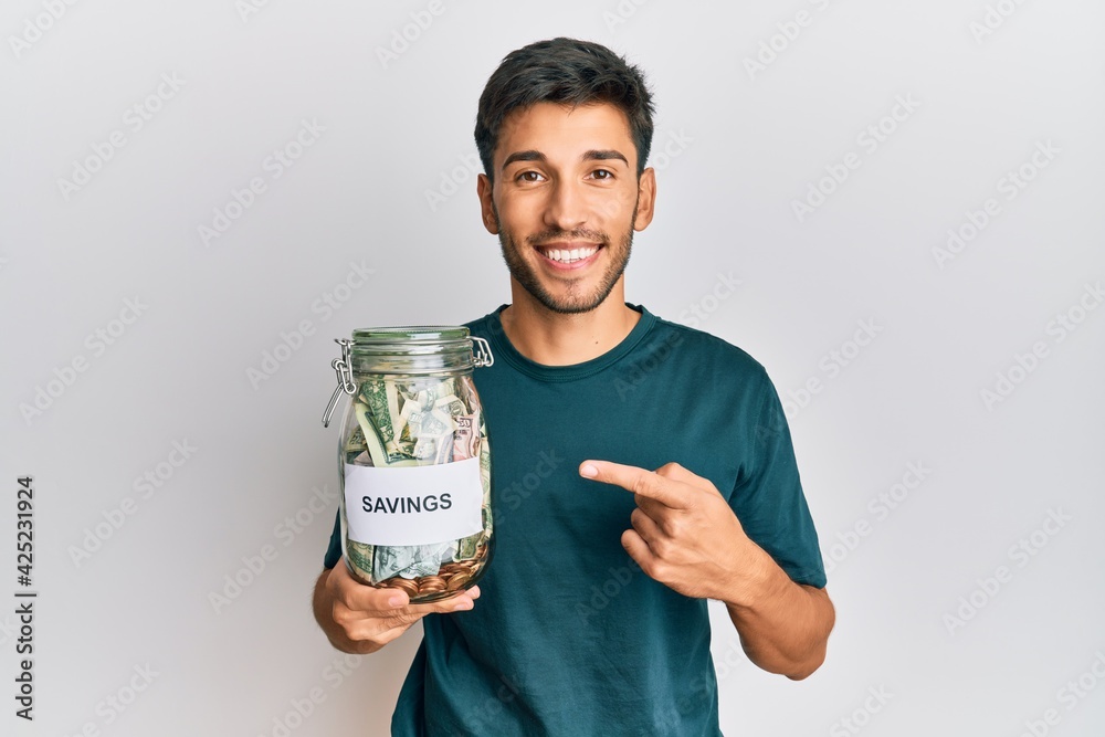 Young handsome man holding jar with savings smiling happy pointing with hand and finger Young handsome man holding jar with savings smiling happy pointing with hand and finger