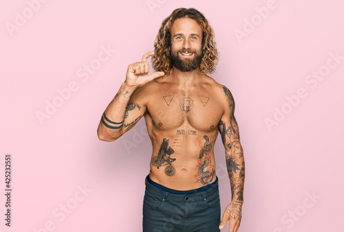 Handsome man with beard and long hair standing shirtless showing tattoos smiling and confident gesturing with hand doing small size sign with fingers looking and the camera. measure concept.