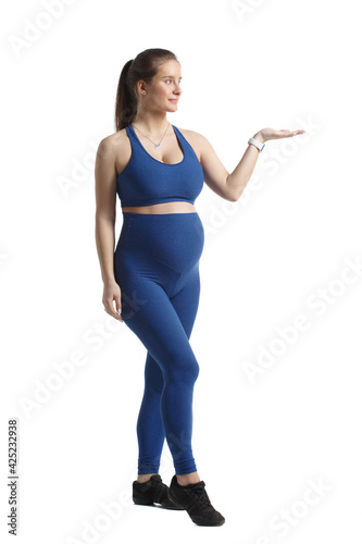 Fototapeta Naklejka Na Ścianę i Meble -  Cute pregnant woman in blue sportswear stands in full growth and points with her hand to the right isolated on a white background.